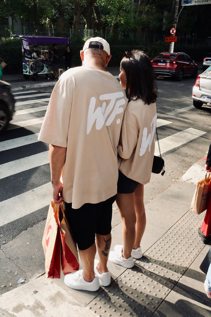 Couple in matching shirts stand at a crosswalk in a sunny urban setting.
